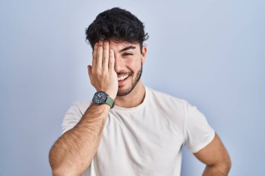 Beyaz arka planda duran İspanyol sakallı bir adam bir gözünü eliyle kapatıyor, yüzünde kendine güvenen bir gülümseme ve sürpriz bir duygu var.. 