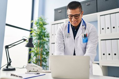 Young latin man doctor using laptop standing at clinic
