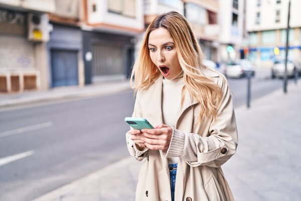 Молодая блондинка, использующая смартфон с неожиданным выражением лица на улице