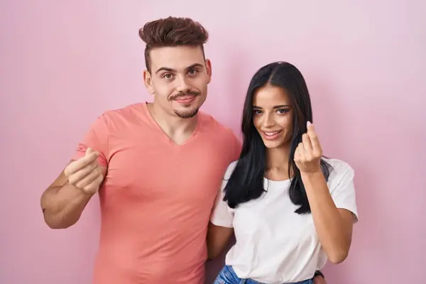 Young Hispanic Couple Standing Pink Background Doing Money Gesture Hands —  Fotos de Stock