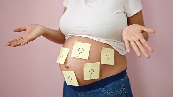 Young pregnant woman with question marks on belly and i dont know gesture over isolated pink background