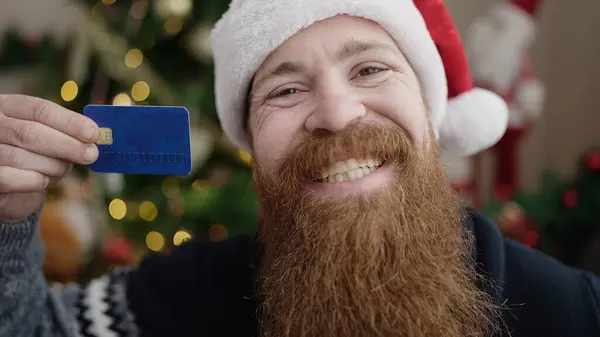 Young redhead man holding credit card standing by christmas tree at home