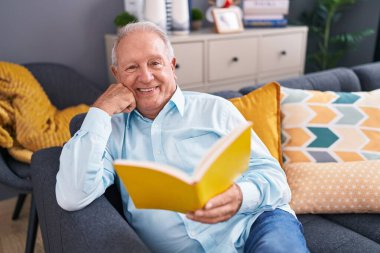 Orta yaşlı, kır saçlı, kitap okuyan adam evdeki kanepede oturuyor.