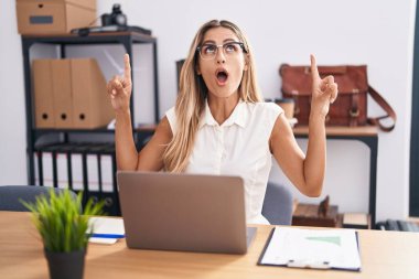 Ofiste çalışan genç sarışın kadın şaşırmış ve şaşırmış bir şekilde yukarı bakıyor ve parmaklarıyla işaret ediyor ve kollarını kaldırıyordu.. 