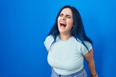 Young modern girl with blue hair standing over blue background angry and mad screaming frustrated and furious, shouting with anger. rage and aggressive concept. 