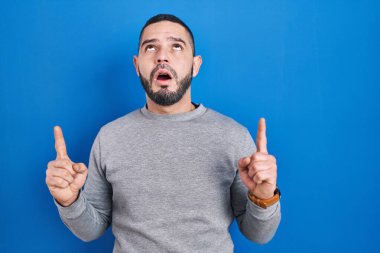 Hispanic man standing over blue background amazed and surprised looking up and pointing with fingers and raised arms. 