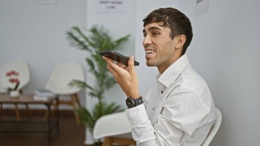 Genç İspanyol adam, bekleme odasındaki sandalyede oturan akıllı telefonuyla sesli mesaj gönderiyor.