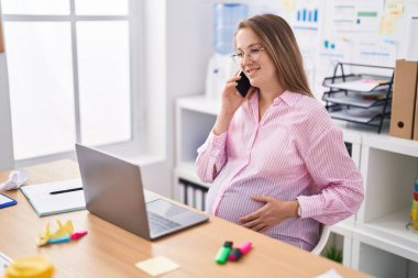 Genç hamile iş kadını bilgisayarını kullanarak ofisteki akıllı telefondan konuşuyor.
