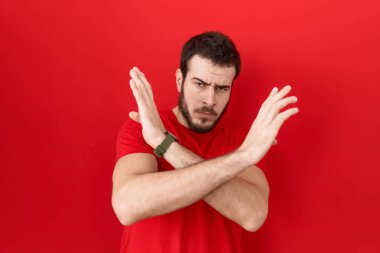 Latin asıllı genç adam, günlük kırmızı tişört giyiyor. Reddedilmiş yüz ifadesi, kollarını kavuşturarak negatif işaret, kızgın yüz ifadesi yapıyor. 