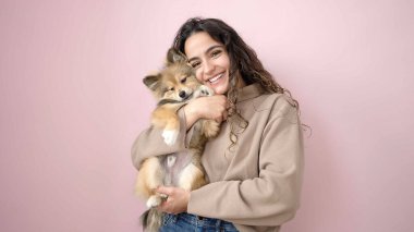 Köpeğiyle gülümseyen genç İspanyol kadın pembe arka planda duruyor.