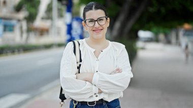 Genç, güzel, İspanyol bir kadın gülümsüyor caddede kollarını kavuşturmuş duruyor.