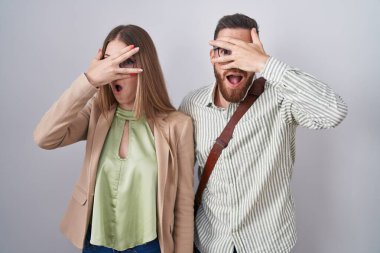 Beyaz arka planda duran genç çift şok içinde yüzlerini ve gözlerini kapayarak utangaç ifadesiyle parmaklarının arasından bakıyor.. 