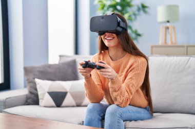 Evde sanal gerçeklik gözlüğü ve joystick kullanan güzel İspanyol bir kadın oyun oynuyor.