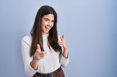 Mavi arka planda duran esmer kadın mutlu ve komik yüzlü parmaklarını kameraya doğrultuyor. iyi enerji ve titreşimler. 