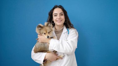 Köpek veterineriyle gülümseyen genç İspanyol kadın izole edilmiş mavi arka planda sarılıyor.