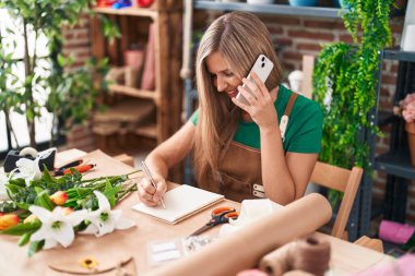 Young blonde woman florist talking on smartphone writing on notebook at flower shop