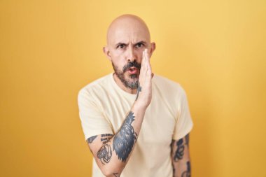 Sarı arka planda dikilen İspanyol dövmeli adam ağzının üstünde gizli dedikodular söylüyor, kötü niyetli konuşmalar fısıldıyor. 