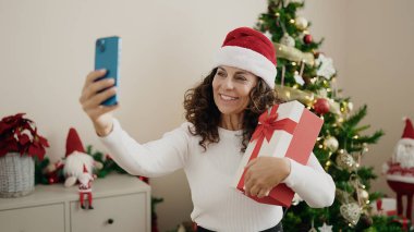 Orta yaşlı İspanyol kadın evde elinde Noel hediyesi olan akıllı telefonuyla selfie çekiyor.