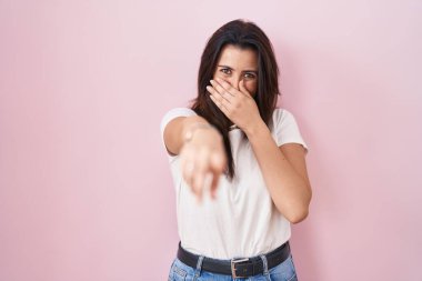 Genç esmer kadın pembe arka planda durup sana gülüyor, eli ağzında kamerayı işaret ediyor, yüz ifadesi utanç verici. 