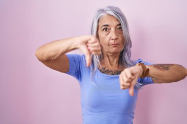 Pembe arka planda dövmeleri olan orta yaşlı bir kadın baş parmaklarını kaldırıyor, anlaşmazlık ve anlaşma ifadesi takınıyor. çılgın çatışma 