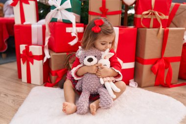 Sevimli sarışın bebek elinde oyuncak ayıyla evde Noel hediyeleriyle oturuyor.