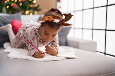 Adorable hispanic girl drawing on notebook lying on sofa by christmas tree at home