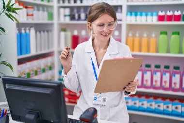 Young blonde woman pharmacist using computer reading document at pharmacy