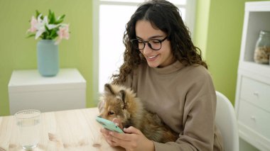 Yemek odasında akıllı telefon kullanan köpekli genç İspanyol kadın.