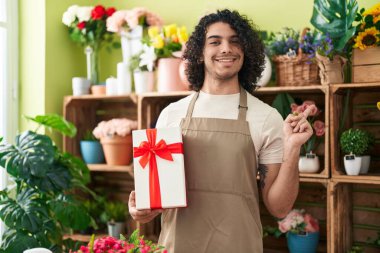 Çiçekçi dükkanında çalışan kıvırcık saçlı İspanyol bir adam elinde hediyeyle mutlu bir şekilde gülümsüyor. 