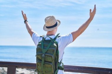 Young caucasian man tourist standing with arms open on back view at seaside