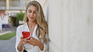 Akıllı telefonuna dalmış şık, genç sarışın bir kadın, güneşli bir şehir arka planında duruyor, teknoloji meraklısı şehir yaşamını örnek alıyor..