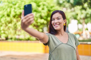 Genç, güzel, İspanyol bir kadın gülümsüyor. Parktaki akıllı telefonun yanında özçekim yapıyor.