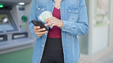 Şili pesolarını elinde tutan genç, güzel İspanyol kadın banka veznedarında akıllı telefon kullanıyor.