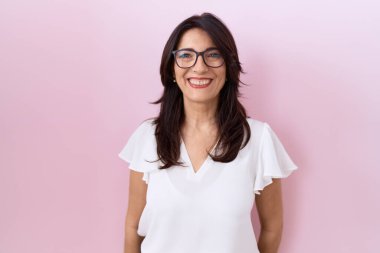 Orta yaşlı İspanyol kadın, günlük beyaz tişört ve gözlük takıyor ve yüzünde mutlu ve havalı bir gülümseme var. şanslı kişi. 