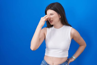 Young caucasian woman standing over blue background smelling something stinky and disgusting, intolerable smell, holding breath with fingers on nose. bad smell 