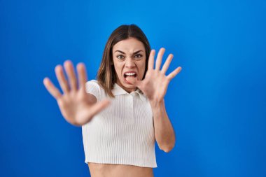 Mavi arka planda duran İspanyol kadın korkudan dehşete düşmüş ve korkmuş. El hareketlerini durdur, şok içinde bağır. panik konsepti. 