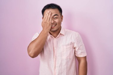 Çinli genç adam pembe arka planda duruyor. Bir gözünü eliyle kapatıyor. Yüzünde kendine güvenen bir gülümseme ve sürpriz bir duygu var.. 