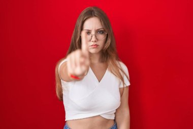 Kırmızı arka planda dikilen genç beyaz kadın parmağını kaldırmış ve kızgın bir ifadeyle hiçbir hareket göstermiyor. 