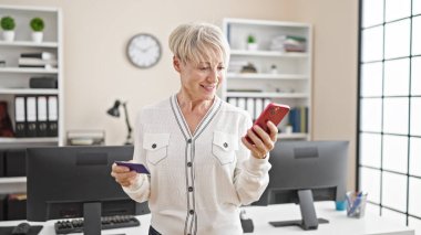 Orta yaşlı sarışın iş kadını. Ofiste akıllı telefon ve kredi kartıyla alışveriş yapıyor.