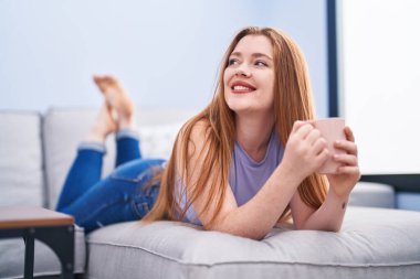 Genç kızıl saçlı kadın evdeki kanepede uzanmış kahve içiyor.