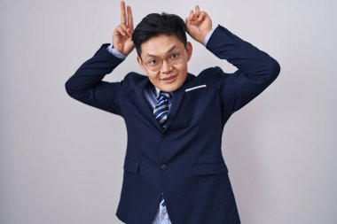 Genç Asyalı takım elbiseli ve kravatlı komik ve çılgın pozlar veren parmakları tavşan kulaklı, neşeli gülümseyen 