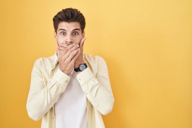 Sarı arka planda duran İspanyol genç adam yanlışlıkla ağzı ellerle kapatarak şok geçirdi. gizli kavram. 
