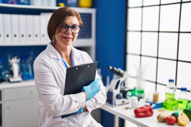 Orta yaşlı kadın bilim adamı gülümsüyor. Laboratuvarda elinde bir pano var.