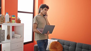 Genç İspanyol adam yemek odasında bilgisayarla akıllı telefondan konuşuyor.