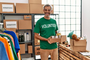 Orta yaşlı, gri saçlı gönüllü, güler yüzlü, hayır kurumunda konserve yiyecek tutan.