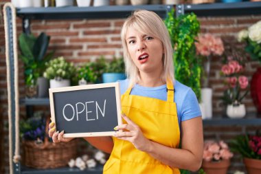 Young caucasian woman working at florist holding open sign clueless and confused expression. doubt concept. 
