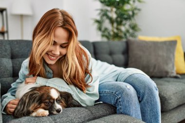 Genç beyaz kadın kanepede uzanmış evde köpeğe sarılıyor.
