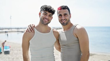 İki erkek, deniz kenarında birbirlerine sarılarak gülümsüyor.