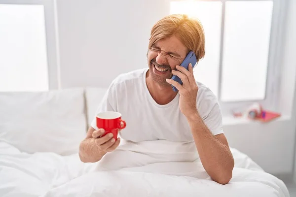 Genç Sarışın Adam Akıllı Telefonda Konuşuyor Yatak Odasında Kahve Içiyor — Stok fotoğraf