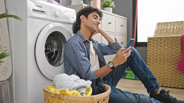 Young hispanic man using smartphone waiting for washing machine at laundry room